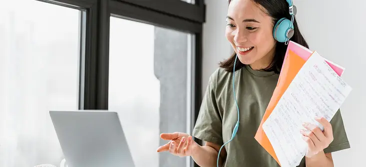 A smiling woman wearing headphones engages in an online conversation while holding notes in front of a laptop