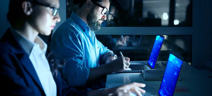 Two professionals work late at night in a dimly lit office, focused on laptops