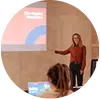 "A woman standing and speaking in front of a group of people in a room with a large screen displaying a rainbow and a cloud."