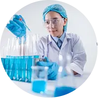 A scientist or lab technician wearing a white lab coat, blue gloves, and a hairnet working with test tubes filled with a blue liquid in a laboratory setting.